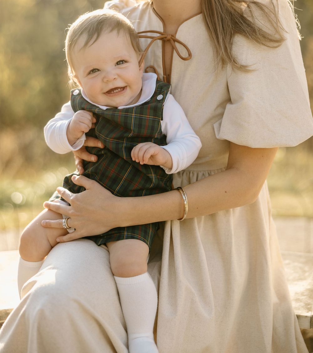 Baby in white shirt and overalls smiles while being held outdoors in warm sunlight.