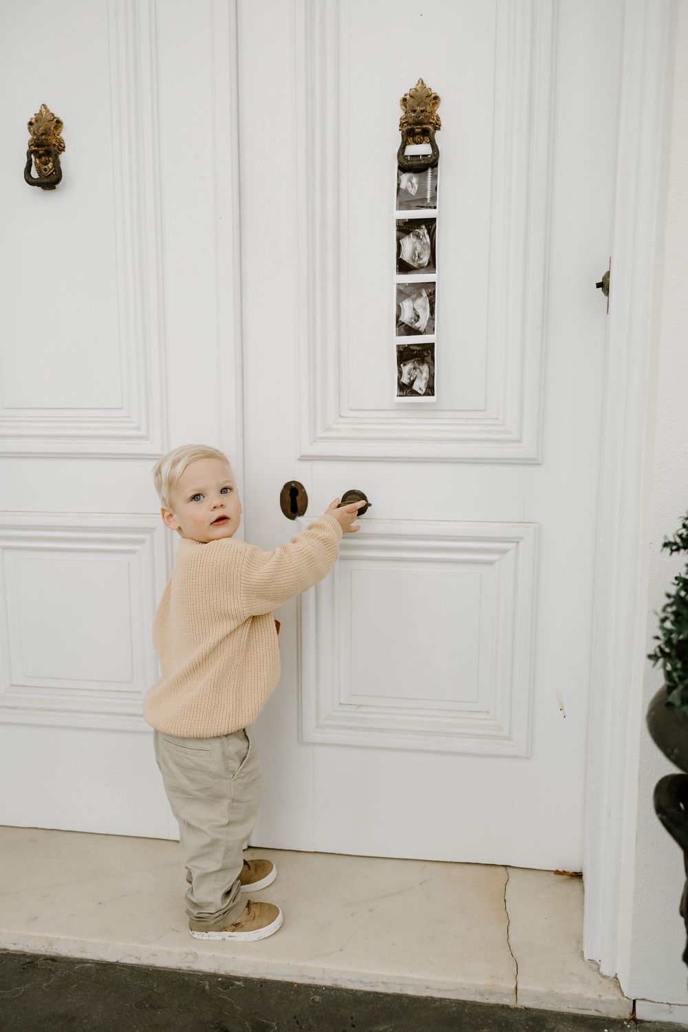 A toddler in beige sweater and khaki pants reaches up to touch decorative wall sconces in a white room.