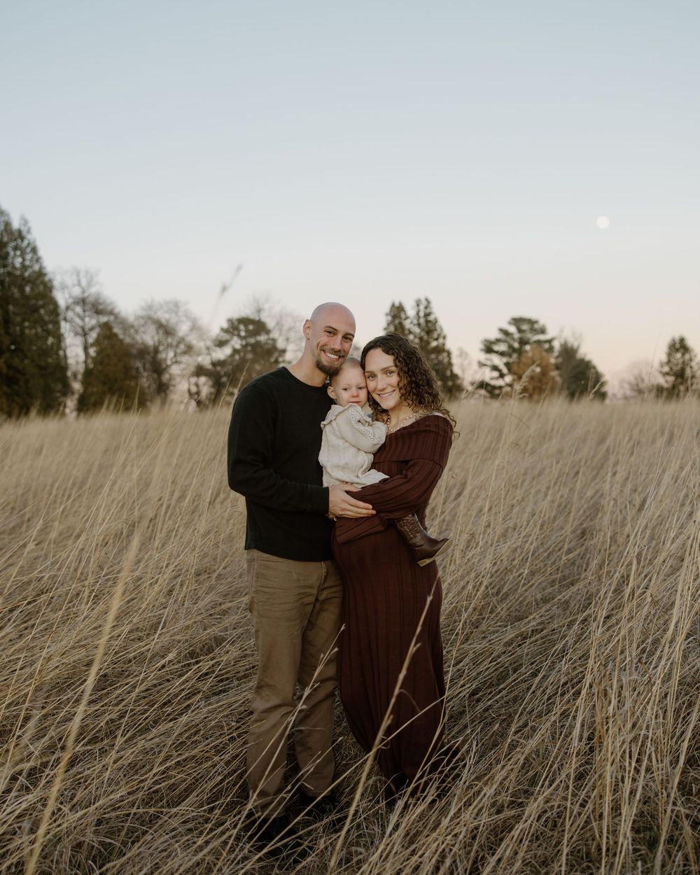 A couple embraces tenderly while standing in a golden field of tall grass at sunset.