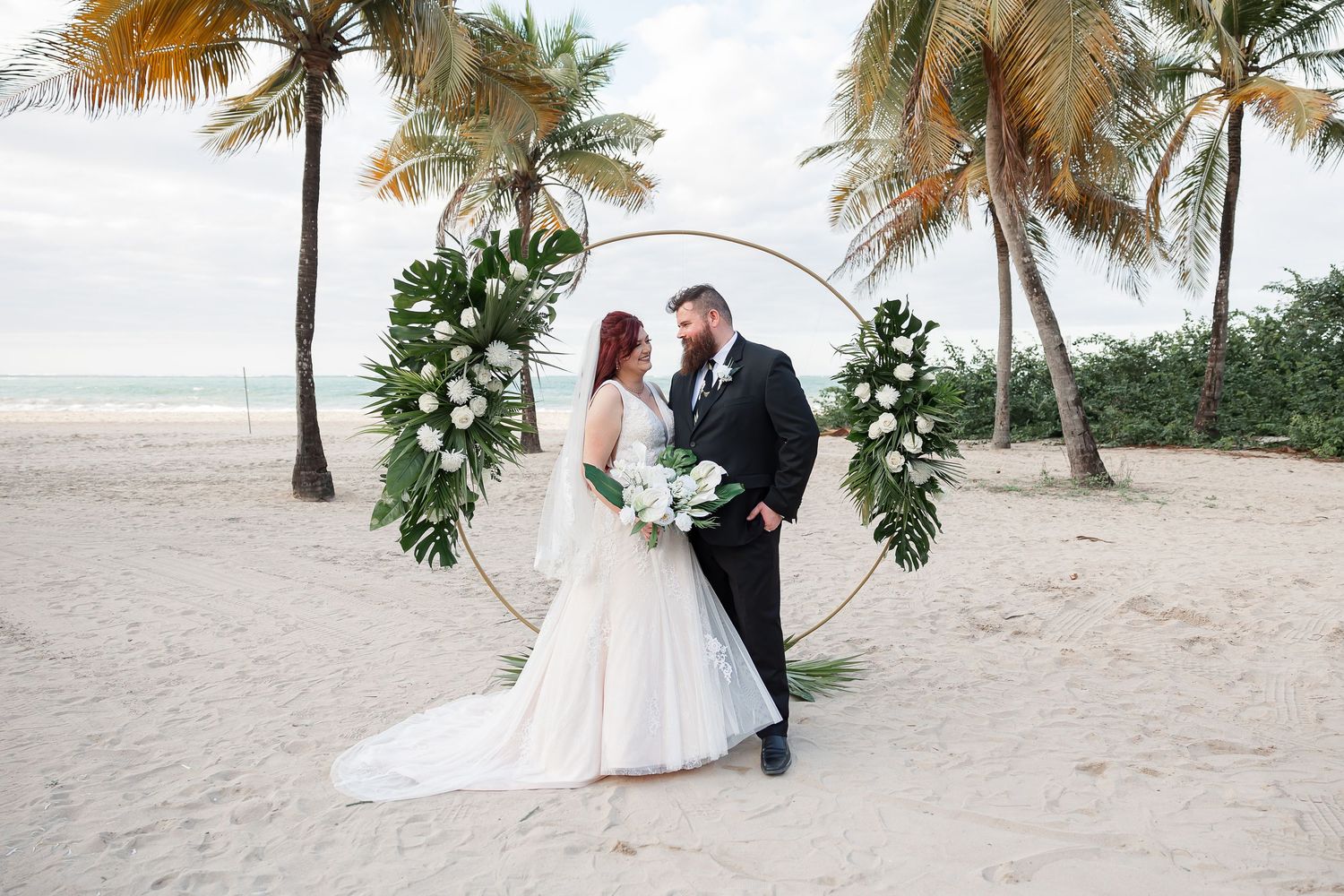 A bride and groom embrace on a beach with palm trees and the ocean in the background.