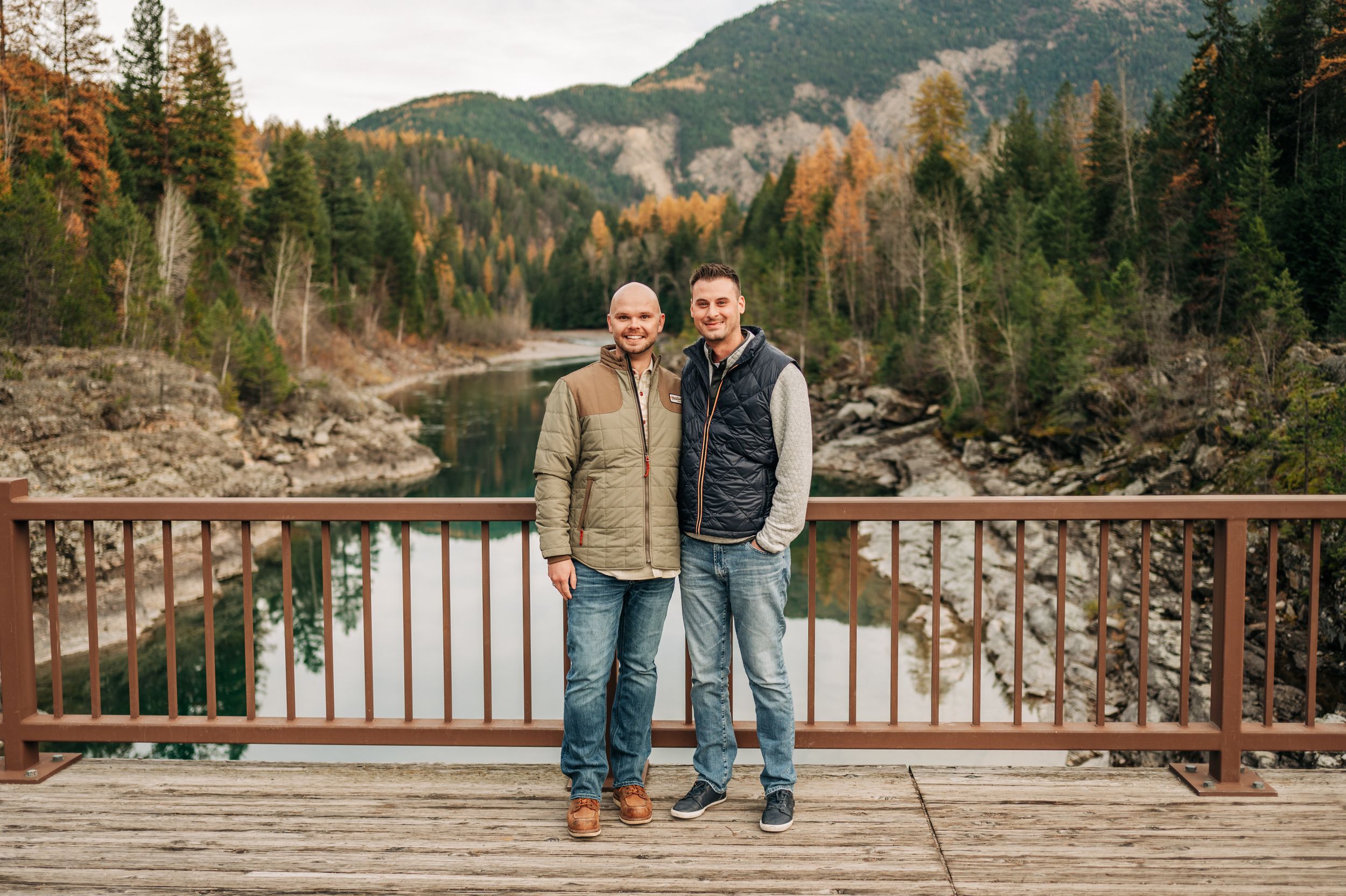 Two people stand on a wooden bridge overlooking a mountain river landscape.