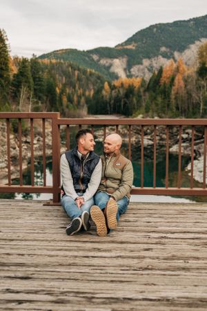 Two people share an intimate moment sitting on a wooden deck overlooking autumn mountains and a lake.