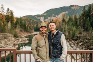 A couple poses together on a scenic bridge deck with fall foliage and mountains in the background.