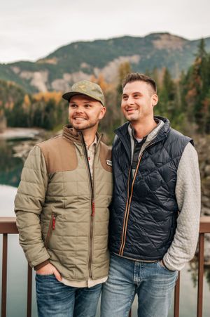 Two people in puffy vests smile while standing on a viewing platform with mountains and water behind them.