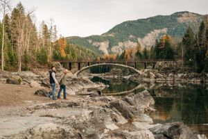 A couple stands by rocky shoreline with historic stone bridge and autumn mountain landscape in background.