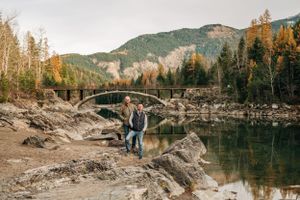 Two people pose near a curved stone bridge with mountains and fall colors reflecting in calm water.