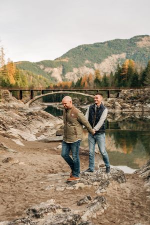 Two people explore rocky terrain near historic bridge with mountain backdrop and autumn trees.