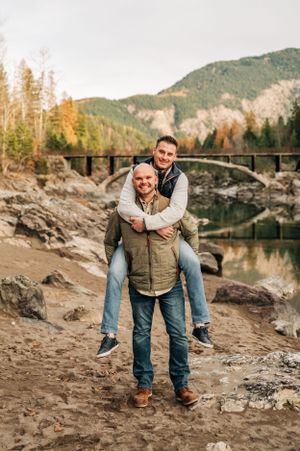 Playful piggyback ride moment captured against mountain landscape and stone bridge background.