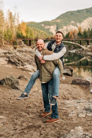 Couple shares playful moment on rocky shore with historic bridge and mountain scenery behind them.