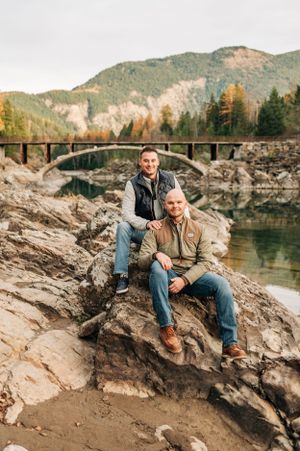 Two people pose on rocky outcrop with stone bridge and mountain vista in background.
