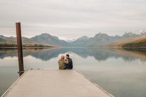 A couple sits at the end of a wooden pier overlooking a peaceful mountain lake at dusk.