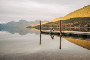 Person sitting on dock with perfect mirror reflection in calm mountain lake waters.