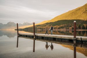 Two figures walking on wooden pier with mountain reflections in still lake waters.