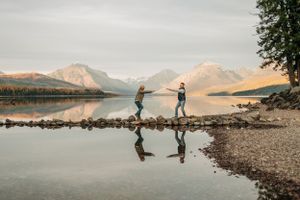 Two people standing on rocky shoreline with perfect reflection in calm mountain lake.