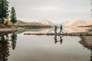 Two silhouettes balanced on rocks by mountain lake with mirror-like reflections at sunset.