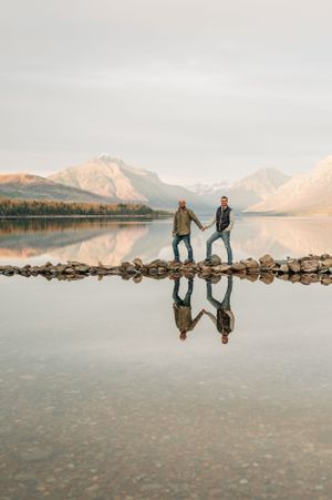 Two people standing on rocky outcrop with perfect reflection in mountain lake at sunset.
