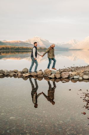 Two people holding hands while balancing on rocks by mountain lake with mirror reflection.