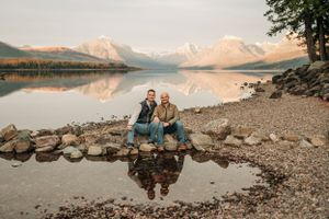 Two people sitting on rocky shore with mountain lake reflections at golden hour.
