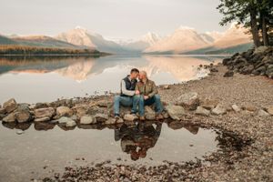 Couple sitting close together on rocky lakeshore with mountains and sunset reflections.