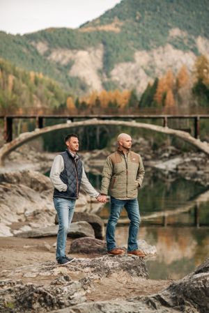 Two people stand on rocky terrain near a stone bridge over a river with autumn mountains in the background.