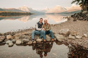 Two people sit on rocky shoreline beside a calm mountain lake with snow-capped peaks reflecting in the water.