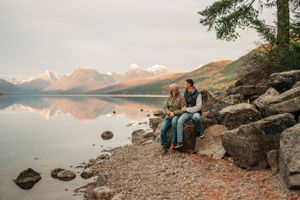 Two people sit together on rocky shore with mountains and lake reflections creating serene landscape.