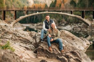 Two people pose on rocky outcrop with stone bridge and autumn foliage in background.