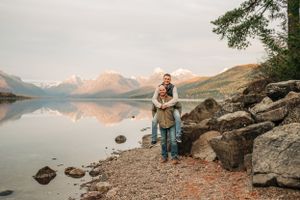 Two people embrace on rocky lakeshore with mountain reflections in calm water at sunset.