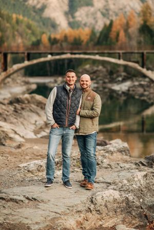 Two people in casual attire stand on rocky terrain with stone bridge and autumn scenery behind them.
