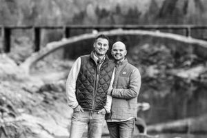 Black and white portrait of two people standing by stone bridge in mountain landscape.