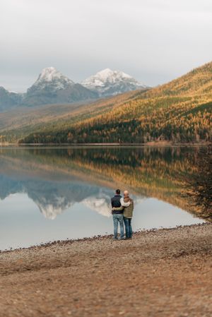 Two people stand at mountain lake shore with snow-capped peaks reflecting in calm water.