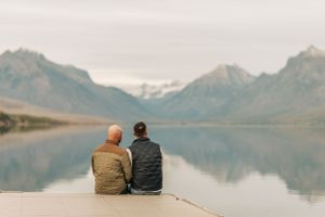 Two people sit together overlooking serene mountain lake with snow-capped peaks in distance.