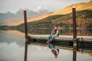 Person sits on wooden dock overlooking mountain lake with autumn colors on distant slopes.