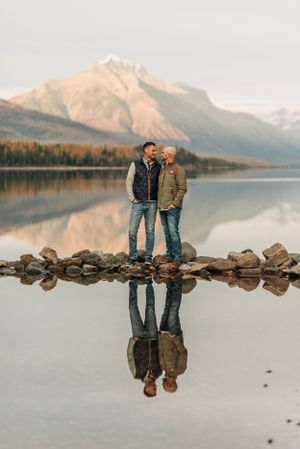 Two people standing on rocky shoreline with mountain reflections in calm lake at sunset.