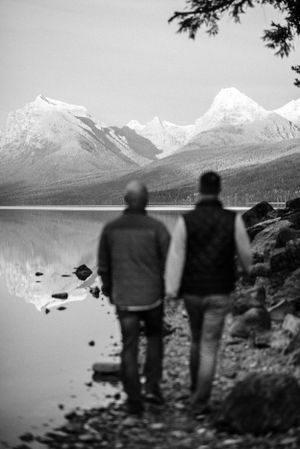 Black and white photo of two silhouettes viewing snow-capped mountains across serene lake.