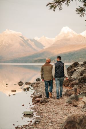 Couple walking along rocky lakeshore with golden mountains in background during sunset.