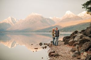 Two people enjoying scenic mountain views while standing on rocky lake shore at golden hour.