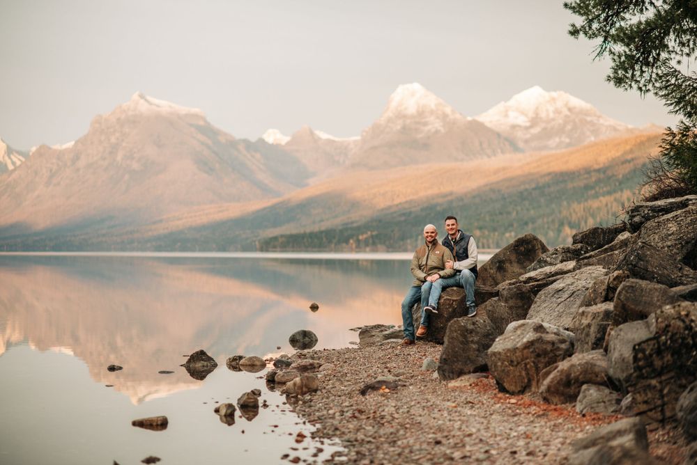 Pair sitting on rocky shoreline admiring mountain reflections in calm lake waters.