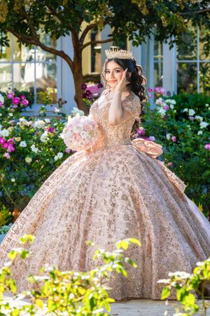 A stunning rose gold ball gown with intricate details sparkles in a garden setting surrounded by blooming flowers.