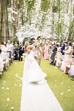 Wedding guests celebrate along a white aisle runner surrounded by bamboo trees as flower petals float through the air.