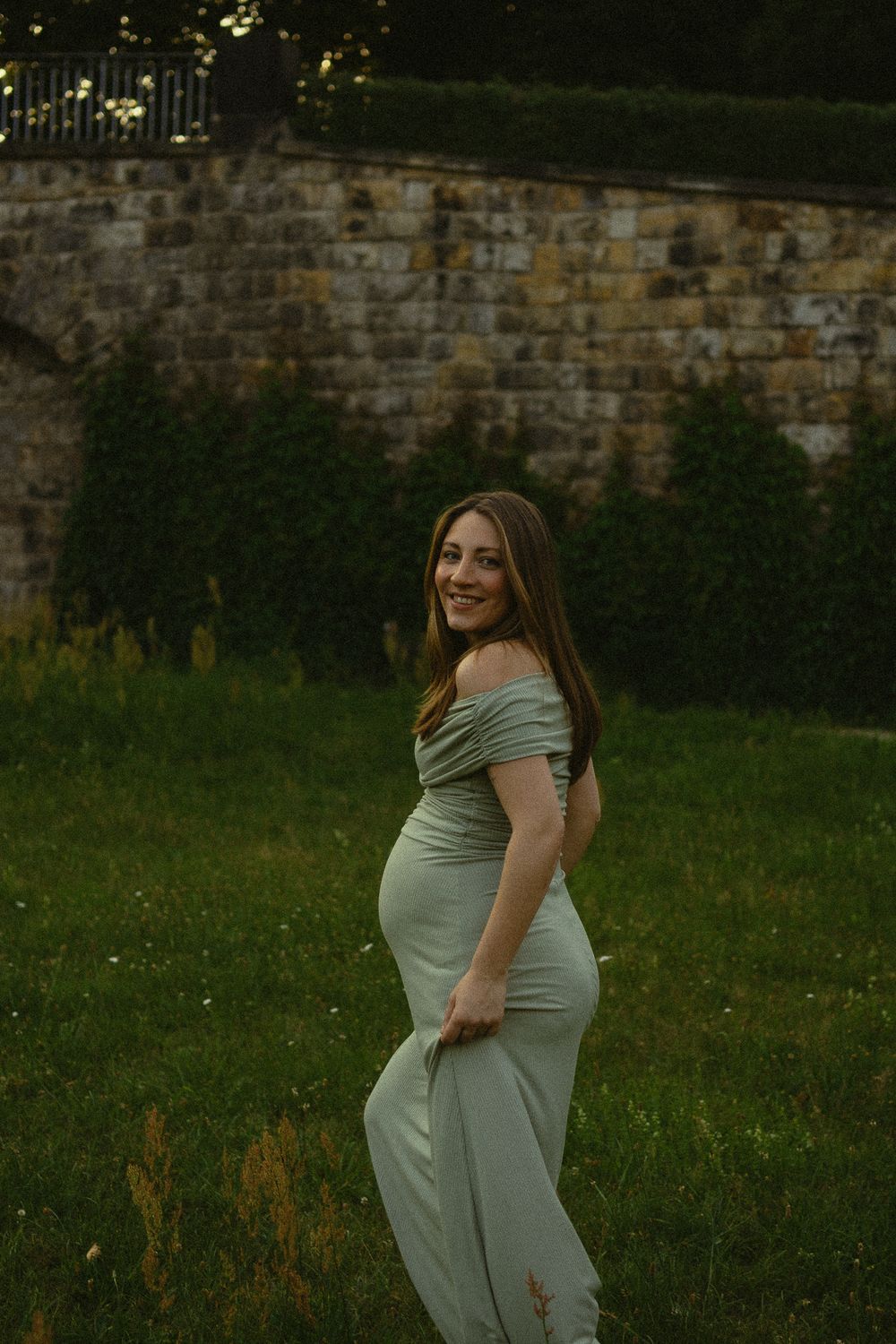 Frau mit Babybauch hält ihr langes grünes Kleid fest und spaziert über Wiese im Park -  Portrait Fotoshooting im Japanischen Palais Dresden