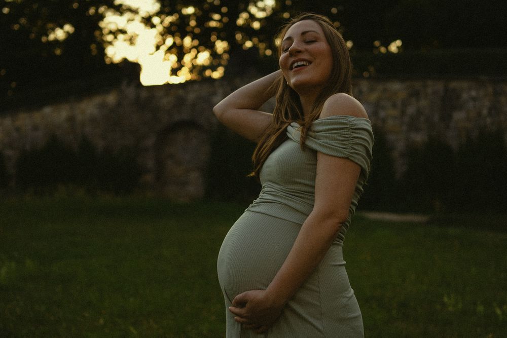 Lachende Frau hält sich ihren Babybauch und streift mit anderer Hand durch Haare - Portrait Fotoshooting bei Sonnenuntergang Dresden