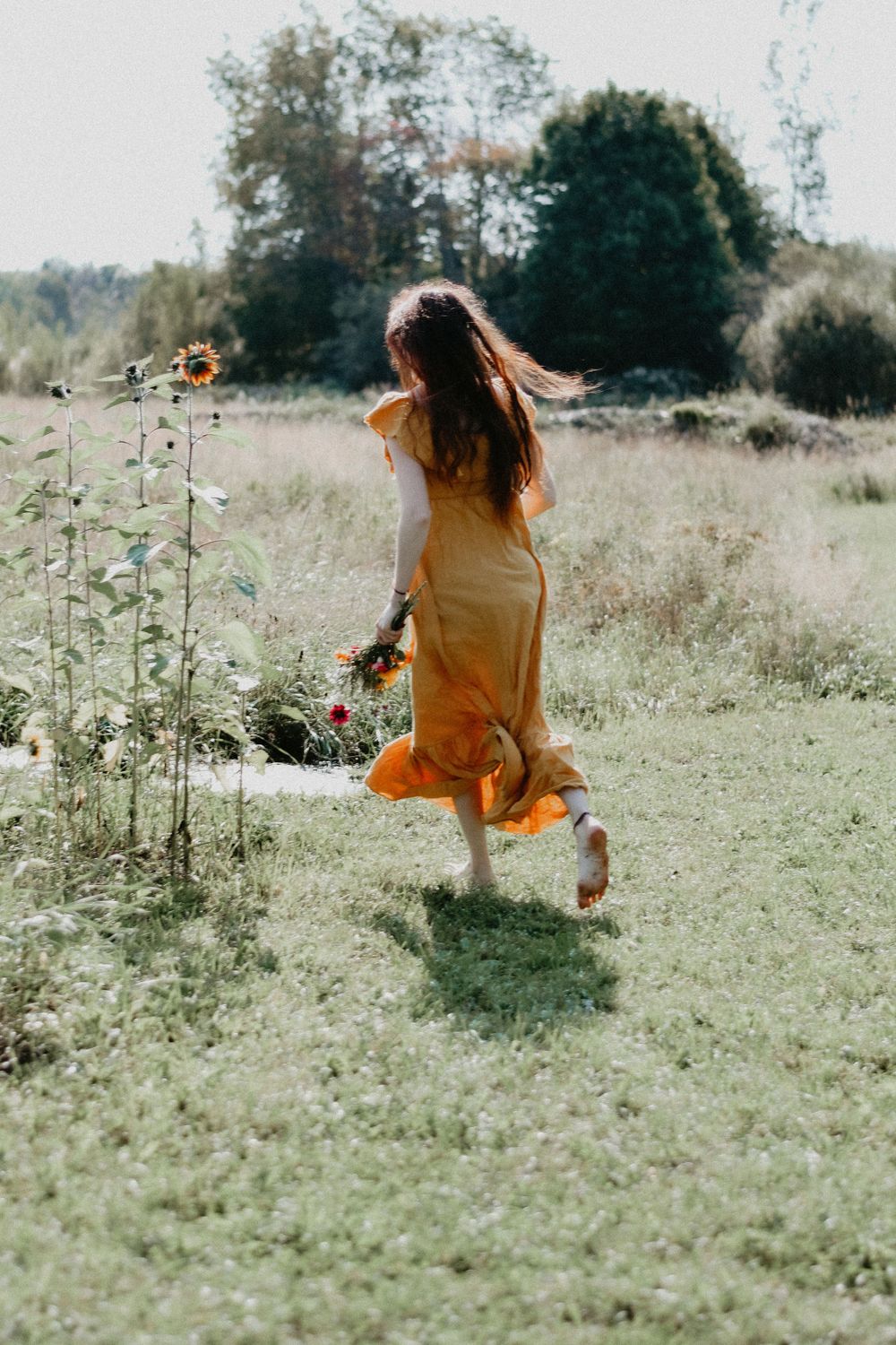A woman in a flowing yellow dress dances through a sunlit meadow near wildflowers on a summer day.