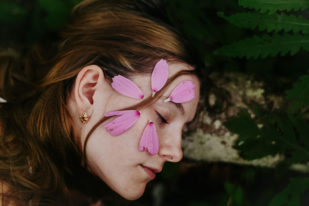 Pink flower petals arranged on a resting person's face against a dark green fern background.