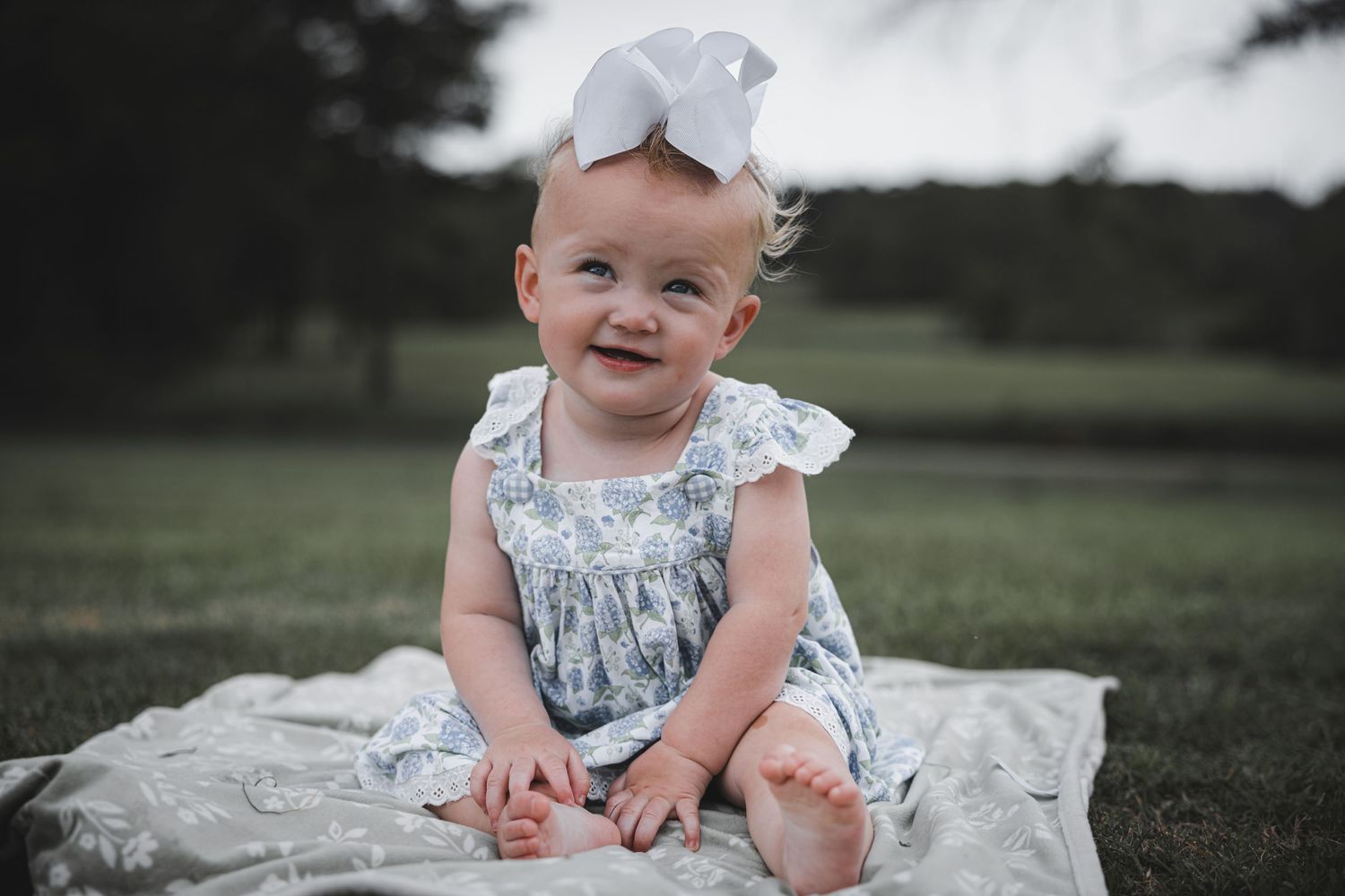 Baby wearing light blue dress and bow sitting on blanket outdoors.