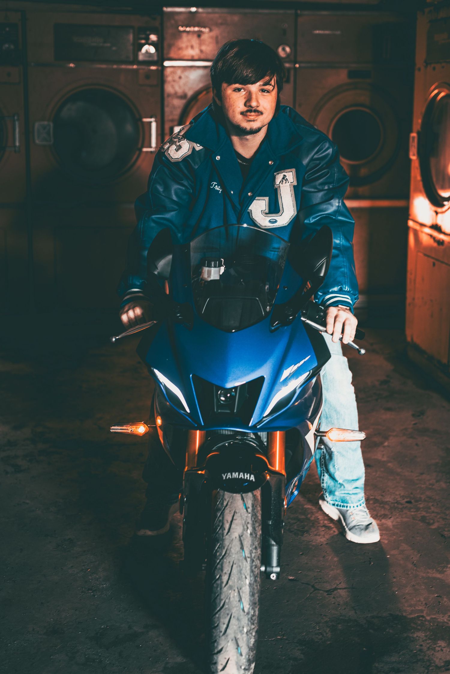 Person on blue Yamaha motorcycle wearing denim jacket in dimly lit garage setting.