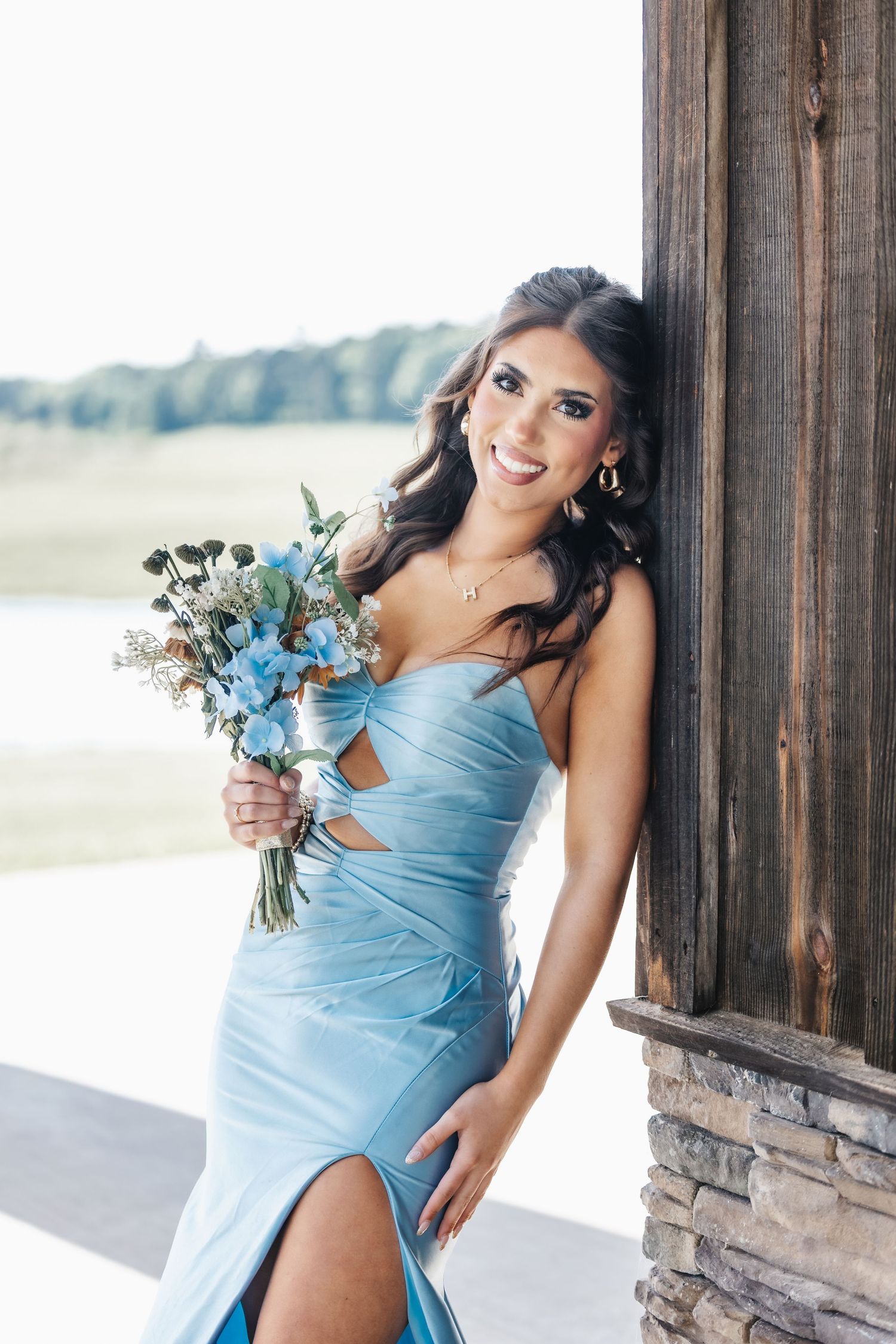 Woman in light blue formal dress holding bouquet posing by wooden column.