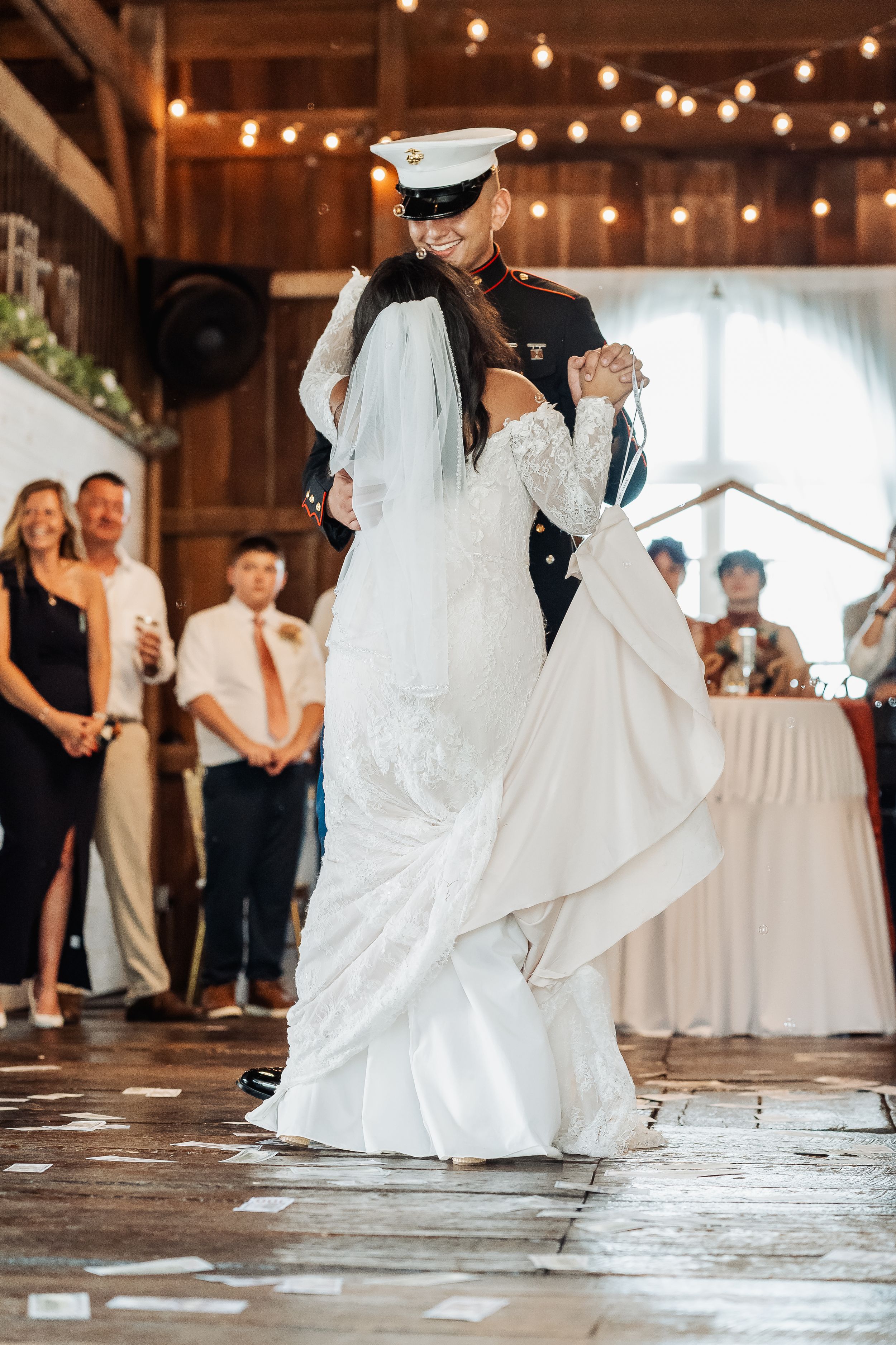 Marine groom lifts bride in white lace wedding dress during first dance in rustic barn venue with string lights.