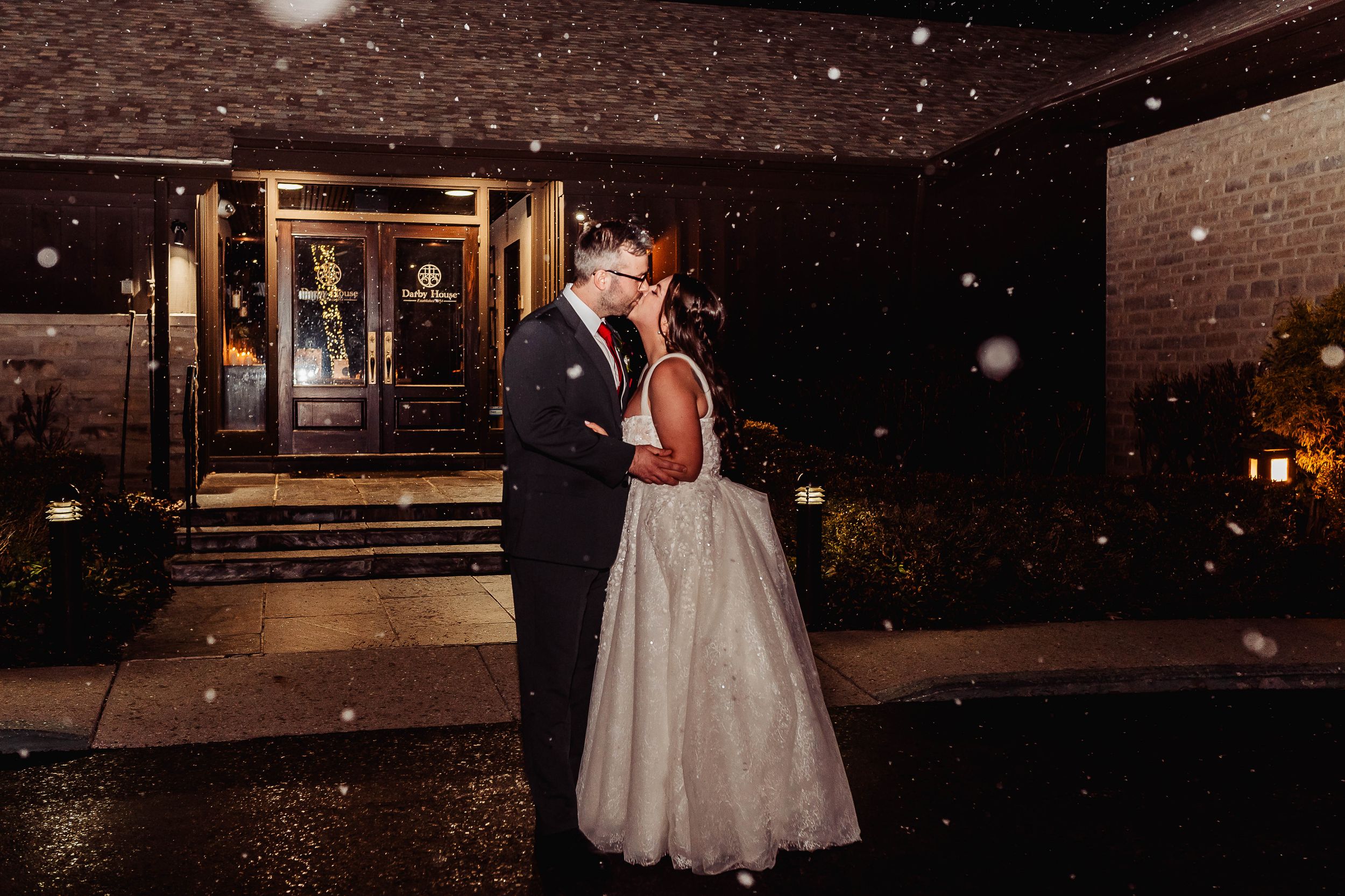 Bride and groom share a romantic kiss outside a venue entrance on a snowy winter wedding night.