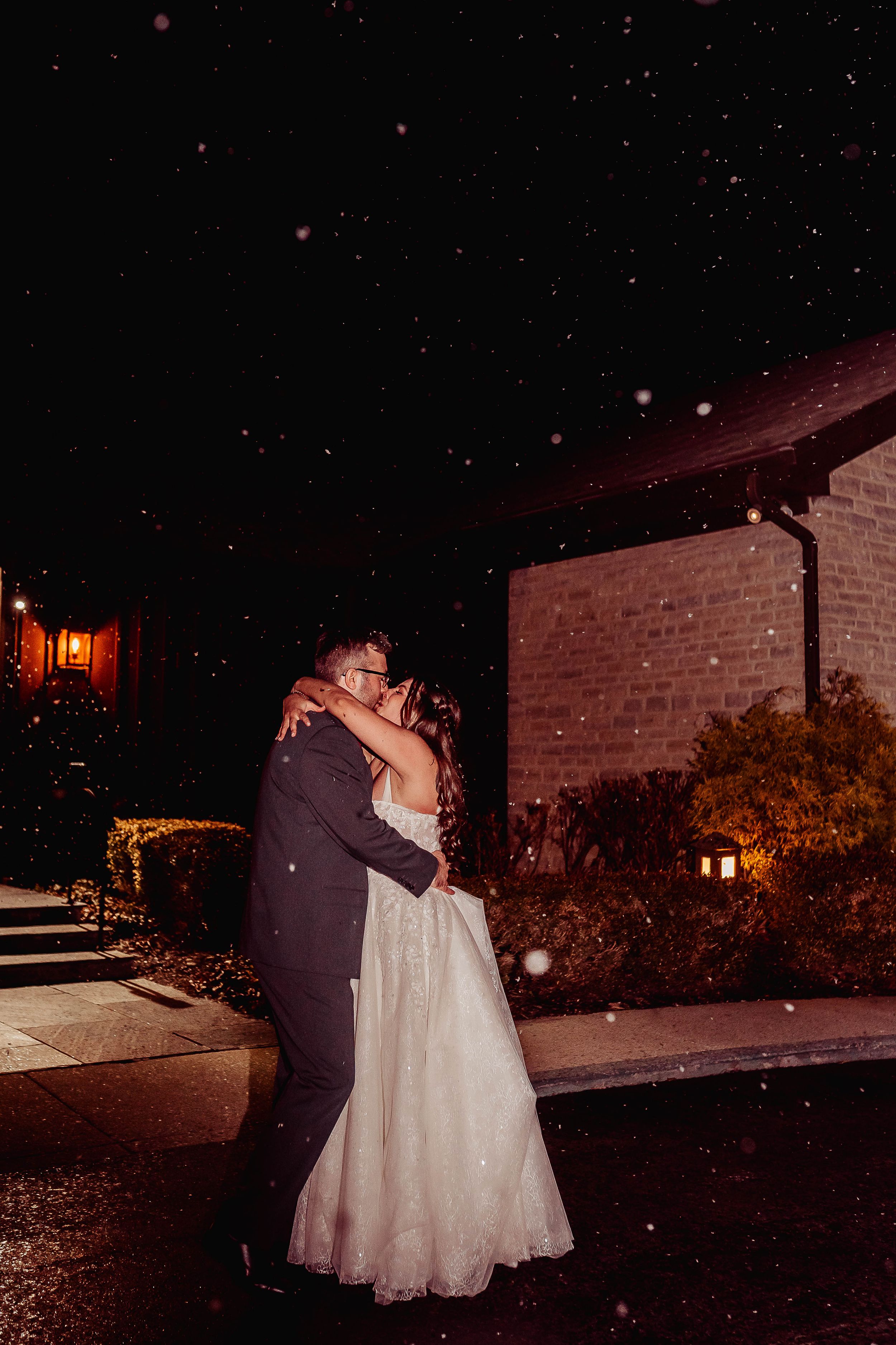 Bride and groom share a romantic kiss outdoors at night in the snow, dressed in wedding attire near a warmly lit venue.